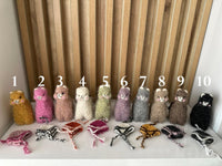 Ten small felted yarn alpaca figures in different colors are lined up on a white surface in front of a wooden slatted backdrop, with white numbers 1 through 10 overlaid above them. Several small woven hats are placed in front of each alpaca.