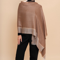 A woman modeling a large taupe-brown shawl with a lighter beige border, styled over a black turtleneck and black trousers against a solid beige studio background.