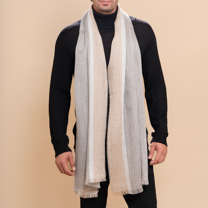 A man modeling a light beige and white striped sateen scarf draped over a black turtleneck sweater and black pants, set against a solid beige studio background.
