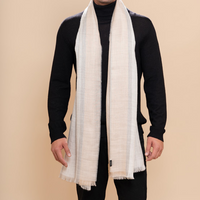 A man modeling a light beige woven scarf with fringed edges, draped over a black turtleneck sweater and black pants against a solid beige background.