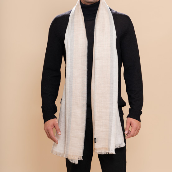 A man modeling a light beige woven scarf with fringed edges, draped over a black turtleneck sweater and black pants against a solid beige background.