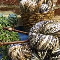Balls of speckled brown and white yarn placed next to a basket and a plant, with knitting needles resting on top of the yarn.